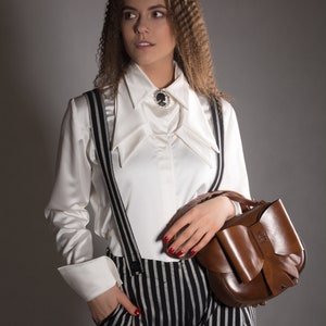 May include: A woman wearing a white collared shirt with a black cameo brooch, black and white striped trousers, and a brown leather handbag with a bow.