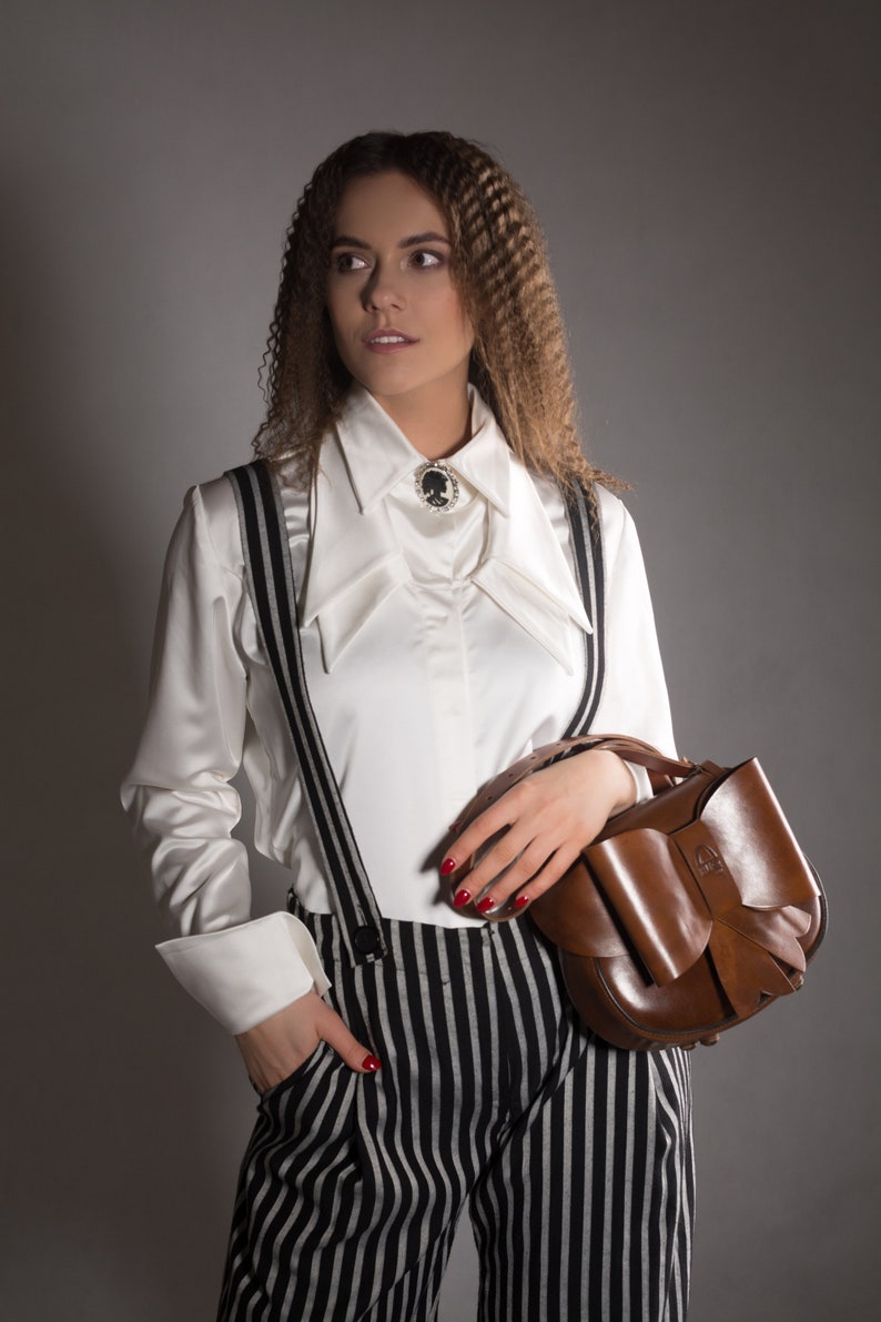 May include: A woman wearing a white collared shirt with a black cameo brooch, black and white striped trousers, and a brown leather handbag with a bow.