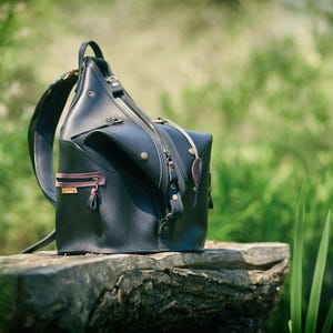 May include: A black leather backpack with brown leather accents and a zipper closure. The backpack is sitting on a wooden log in a grassy area.