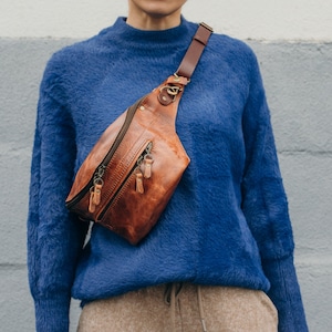 May include: A brown leather fanny pack with a zipper closure and a long strap. The fanny pack is worn over a blue fuzzy sweater.
