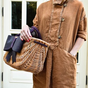 May include: A wicker basket with a large purple leather bow. The basket is being held by a person wearing a brown linen dress.