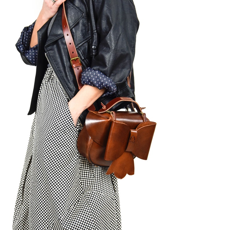 May include: A brown leather satchel bag with a large bow detail. The bag has a long strap and is worn over the shoulder.