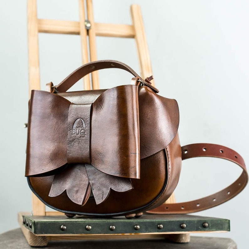 May include: A brown leather handbag with a large bow on the front. The bag has a strap and a buckle. The bag has a logo that says "Lady Bug" on the front.
