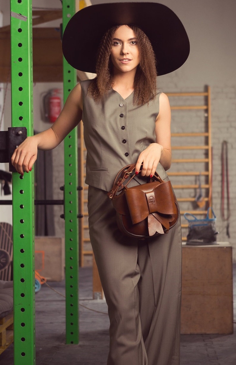 May include: A woman wearing a wide-brimmed black hat and a grey sleeveless vest and trousers. She is holding a brown leather handbag with a large bow.