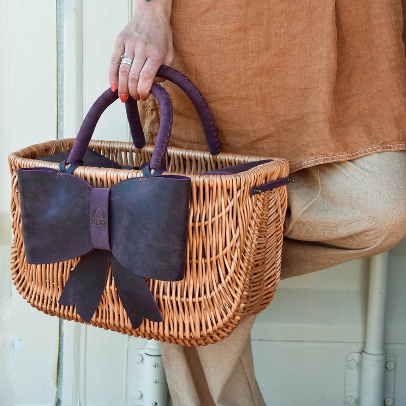 May include: A wicker basket with a purple leather bow and handles. The basket is made of natural woven wicker and has a dark purple leather bow on the front. The handles are also made of leather and are attached to the top of the basket.