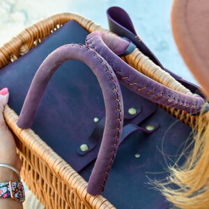 May include: A purple leather tote bag with a woven wicker basket handle. The bag has a flap closure and is accented with brass rivets.