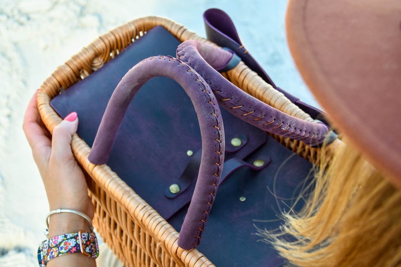 May include: A purple leather tote bag with a woven wicker basket handle. The bag has a flap closure and is accented with brass rivets.