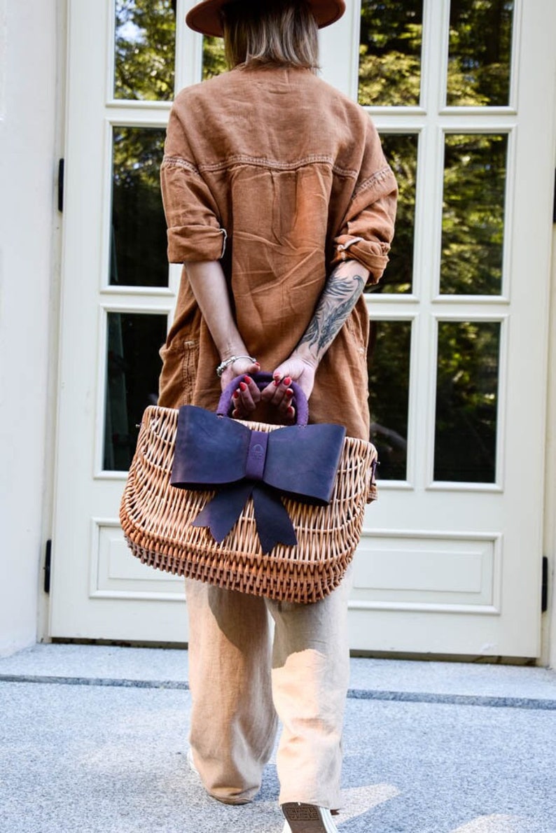 May include: A woman is carrying a woven wicker tote bag with a large purple bow. The bag has a brown leather handle and is made of natural materials.
