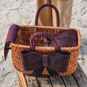May include: A wicker basket with a purple leather bow and handles. The basket is sitting on a wooden surface. The brand name "BUG" is visible on the bow.