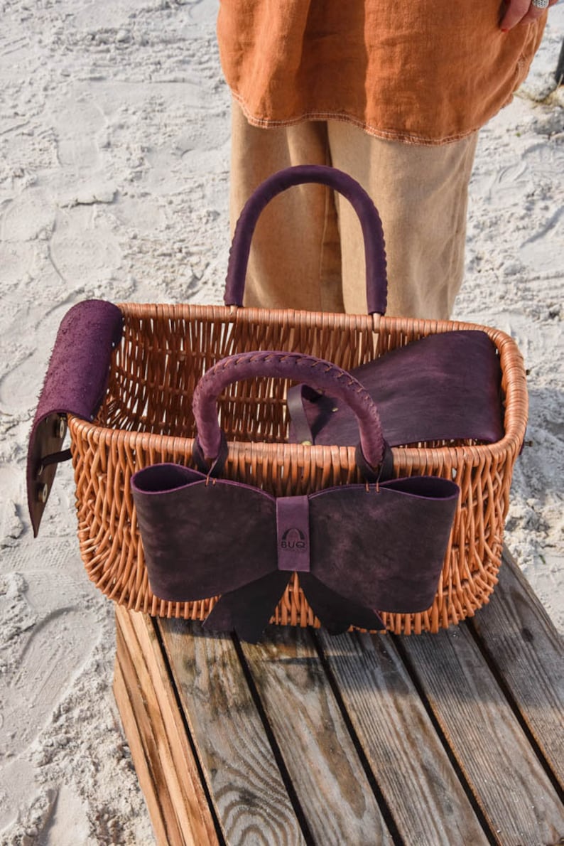 May include: A wicker basket with a purple leather bow and handles. The basket is sitting on a wooden surface. The brand name "BUG" is visible on the bow.