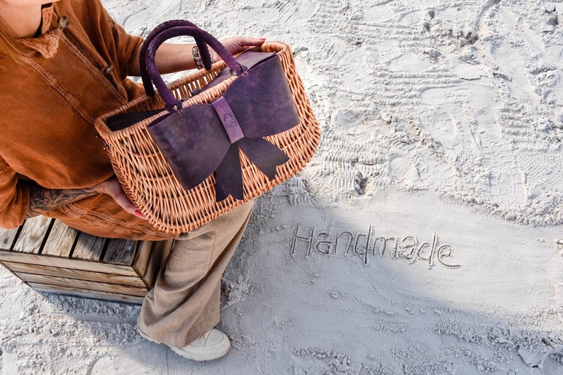 May include: A wicker basket bag with a large purple leather bow. The bag is held by a person wearing a brown jacket and beige pants. The word "Handmade" is written in the sand.