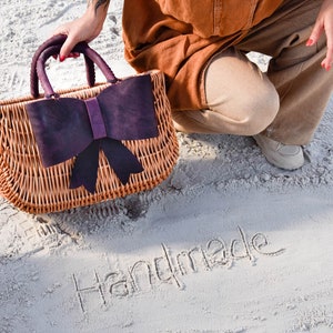 May include: A wicker handbag with a large purple bow on the front. The bag is sitting on white sand with the word "Handmade" written in the sand.