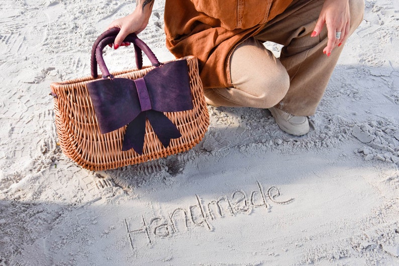 May include: A wicker handbag with a large purple bow on the front. The bag is sitting on white sand with the word "Handmade" written in the sand.