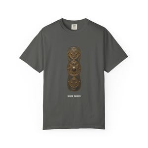May include: A charcoal gray short-sleeve t-shirt featuring a central graphic of an elongated, bronze-colored shield with three circular, ornate embossed patterns. Below the shield, the text RIVER SHIELD is printed in white capital letters.