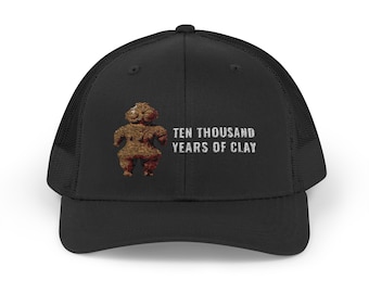 May include: A black trucker hat featuring a mesh back and a solid front panel. The front displays a brown, textured clay figurine graphic positioned to the left of the white, block-lettered text that reads: TEN THOUSAND YEARS OF CLAY.