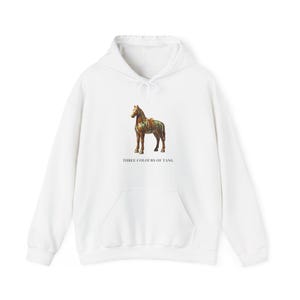 Peut inclure: Un sweat-shirt à capuche blanc avec un motif central représentant une figurine de cheval de la dynastie Tang aux tons marron, vert et jaune. Sous le cheval, le texte THREE COLOURS OF TANG est inscrit en police serif noire.