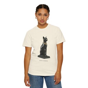 May include: A cream-colored short-sleeve t-shirt featuring a graphic of a black cat statue with a gold collar sitting on a rectangular base. Below the graphic, the text reads QUIET GODDESS in a simple, dark, capitalized font centered on the chest.