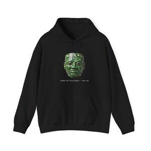 Pode incluir: Uma camisola preta com capuz apresentando um gráfico de uma máscara de mosaico verde no peito. Abaixo da máscara, o texto diz LORD OF PALENQUE // 683 AD. A peça inclui capuz com cordão, mangas compridas e um bolso frontal tipo canguru.