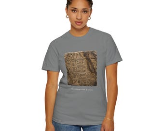 May include: A grey short-sleeve t-shirt featuring a rectangular graphic of ancient Egyptian hieroglyphics carved into stone. Below the graphic, the text "WRITTEN IN STONE & BRASS" is printed in white. The shirt has a crew neckline and a standard fit.