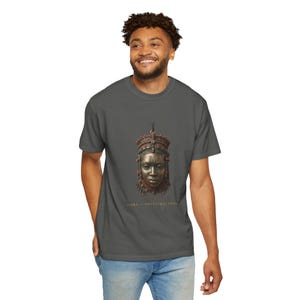 May include: A charcoal gray t-shirt featuring a central graphic of a bronze-toned mask adorned with red beads and intricate patterns. Below the mask, the text reads "YORUBA OF ANCESTRAL GRACE" in gold lettering. The shirt has a crew neckline and short sleeves.