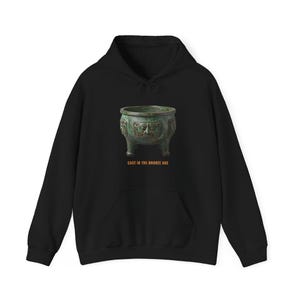 May include: A black hooded sweatshirt featuring a graphic of a green, weathered, three-legged bronze vessel with an embossed face design. Below the vessel, the text CAST IN THE BRONZE AGE is printed in orange capital letters on the chest area.