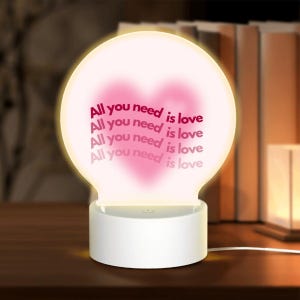 May include: A circular acrylic night light with a white cylindrical base. The light features a soft pink heart graphic and the repeating text "All you need is love" in a dark pink font, illuminated with a warm glow from the base.