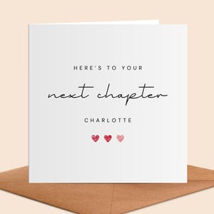 Puede incluir: Una tarjeta de felicitación blanca con el texto "HERE'S TO YOUR next chapter" en escritura negra. Debajo, el nombre "CHARLOTTE" y tres pequeños corazones rojos. La tarjeta está sobre un sobre marrón.