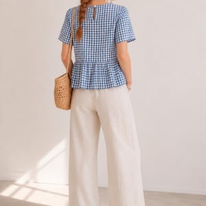 May include: A blue and white gingham short-sleeve peplum top with a round neckline and a button closure at the back, paired with wide-leg off-white trousers and a woven straw shoulder bag. The outfit features a classic check pattern and loose fit.