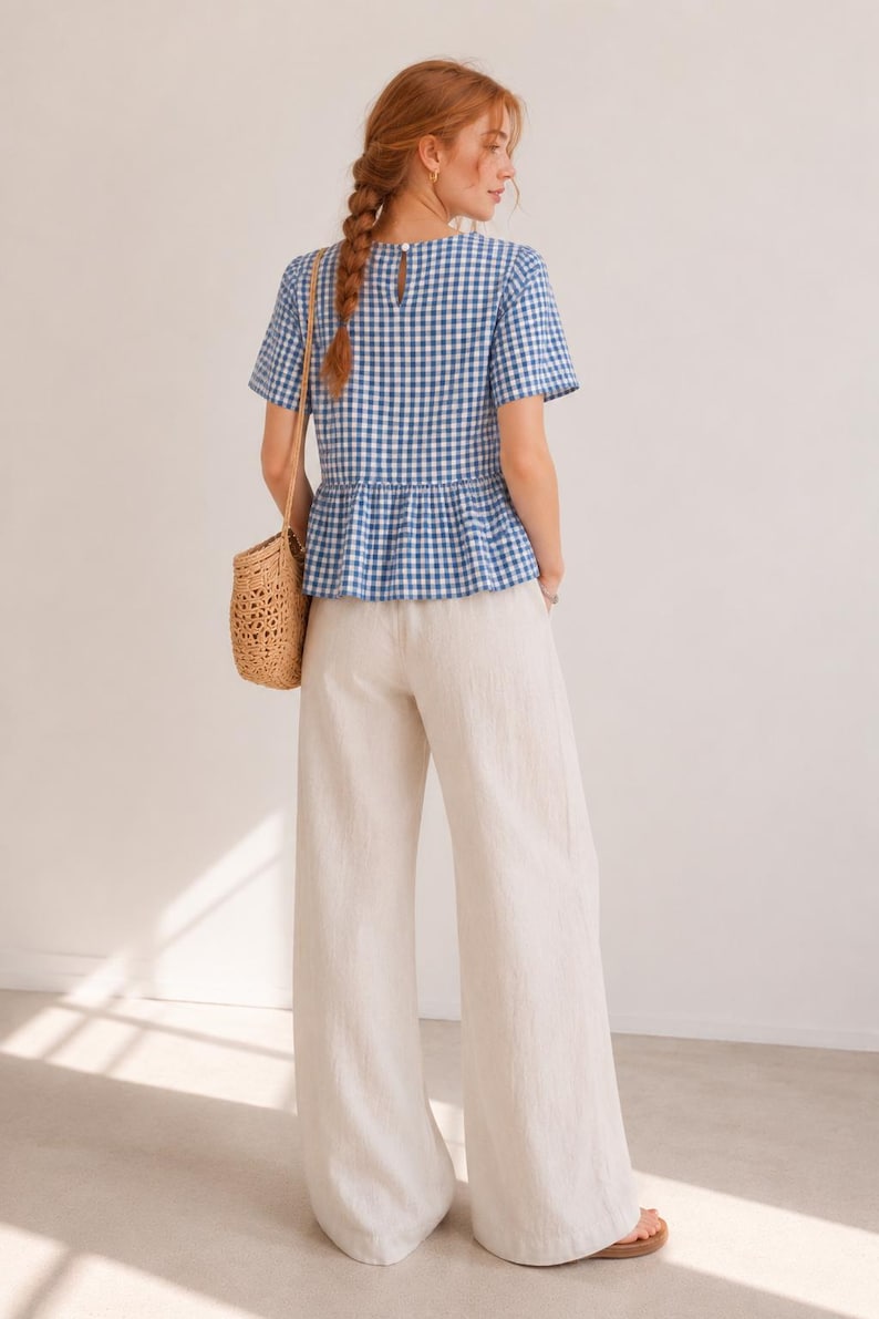 May include: A blue and white gingham short-sleeve peplum top with a round neckline and a button closure at the back, paired with wide-leg off-white trousers and a woven straw shoulder bag. The outfit features a classic check pattern and loose fit.