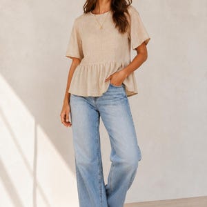 Pode incluir: Um top peplum bege de manga curta com decote redondo e cintura franzida, combinado com cal&ccedil;as de ganga azul-claras de corte largo e design cl&aacute;ssico de cinco bolsos, usado com sand&aacute;lias de dedo castanhas simples.