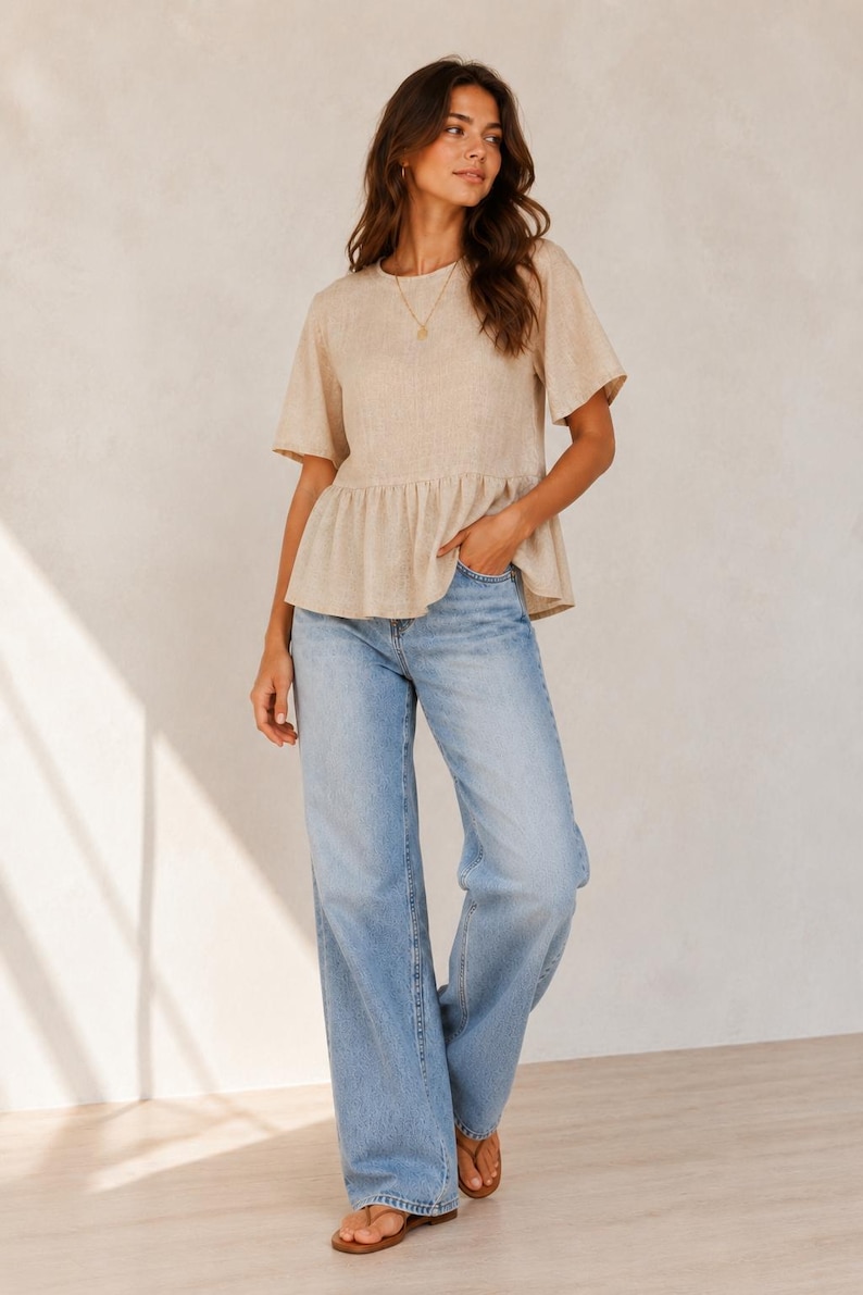 Pode incluir: Um top peplum bege de manga curta com decote redondo e cintura franzida, combinado com cal&ccedil;as de ganga azul-claras de corte largo e design cl&aacute;ssico de cinco bolsos, usado com sand&aacute;lias de dedo castanhas simples.