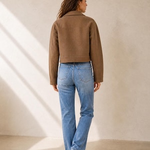 Puede incluir: Una chaqueta corta de color marr&oacute;n con cuello y mangas largas combinada con unos vaqueros de color azul claro con dos bolsillos traseros y corte recto, usados con zapatillas blancas, vistos desde atr&aacute;s para mostrar la silueta completa.