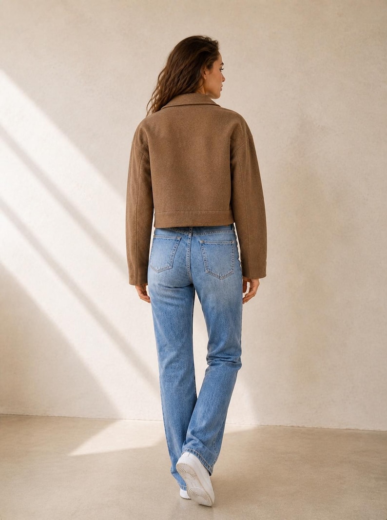 Puede incluir: Una chaqueta corta de color marr&oacute;n con cuello y mangas largas combinada con unos vaqueros de color azul claro con dos bolsillos traseros y corte recto, usados con zapatillas blancas, vistos desde atr&aacute;s para mostrar la silueta completa.