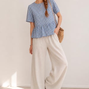 May include: A blue and white gingham print short-sleeved peplum top paired with wide-leg off-white trousers. The top features a round neckline and a gathered waist seam. The trousers have a loose, flowing fit and a simple waistband.