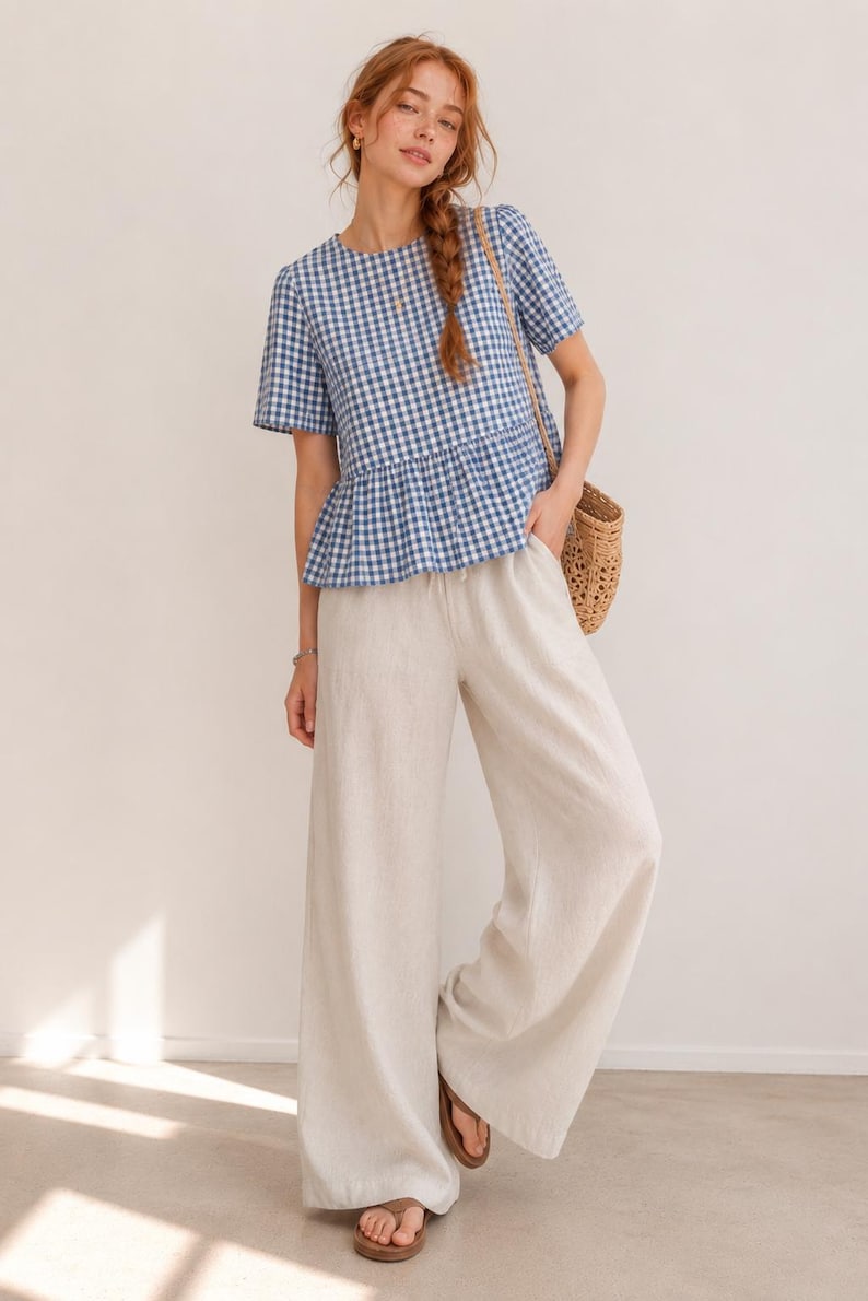 May include: A blue and white gingham print short-sleeved peplum top paired with wide-leg off-white trousers. The top features a round neckline and a gathered waist seam. The trousers have a loose, flowing fit and a simple waistband.