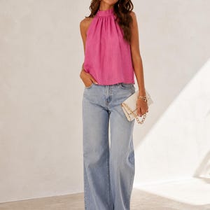 May include: A bright pink sleeveless halter top with a gathered neckline paired with light blue wide-leg denim jeans and a white rectangular clutch bag featuring a gold chain strap. The outfit is completed with open-toe sandals.