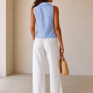 May include: A sleeveless collared top featuring a blue and white vertical striped pattern paired with loose-fitting white wide-leg trousers. The outfit is accessorized with a woven straw handbag with brown handles and simple flat sandals.