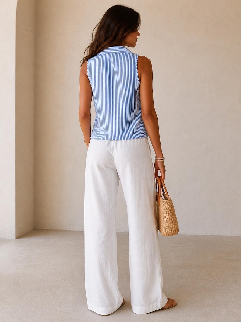 May include: A sleeveless collared top featuring a blue and white vertical striped pattern paired with loose-fitting white wide-leg trousers. The outfit is accessorized with a woven straw handbag with brown handles and simple flat sandals.