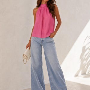 May include: A bright pink sleeveless halter top with a high neckline and gathered fabric detail paired with light blue wide-leg denim jeans and a small white clutch bag with a gold chain strap held in one hand.