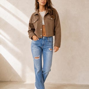 Puede incluir: Una chaqueta corta marr&oacute;n con botones, cuello y dos bolsillos en el pecho, usada sobre un top corto blanco, combinada con jeans azul claro con detalles desgastados en las rodillas y zapatillas blancas.