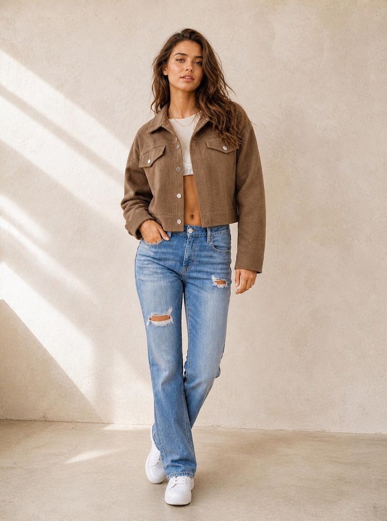 Puede incluir: Una chaqueta corta marr&oacute;n con botones, cuello y dos bolsillos en el pecho, usada sobre un top corto blanco, combinada con jeans azul claro con detalles desgastados en las rodillas y zapatillas blancas.