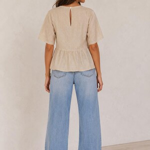 Pode incluir: Um top bege de manga curta com peplum, decote redondo com uma pequena abertura nas costas e fecho de um bot&atilde;o, combinado com cal&ccedil;as de ganga largas em azul claro com um design cl&aacute;ssico de cinco bolsos e bainha reta.