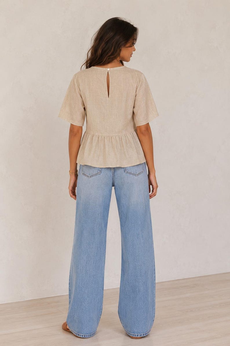 Pode incluir: Um top bege de manga curta com peplum, decote redondo com uma pequena abertura nas costas e fecho de um bot&atilde;o, combinado com cal&ccedil;as de ganga largas em azul claro com um design cl&aacute;ssico de cinco bolsos e bainha reta.