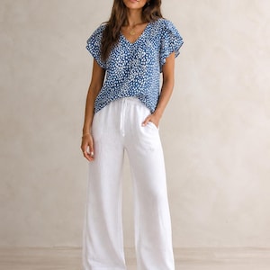 May include: A blue V-neck blouse with a white speckled pattern and flutter sleeves paired with white wide-leg trousers featuring a drawstring waist and side pockets. The outfit is completed with brown open-toe sandals.