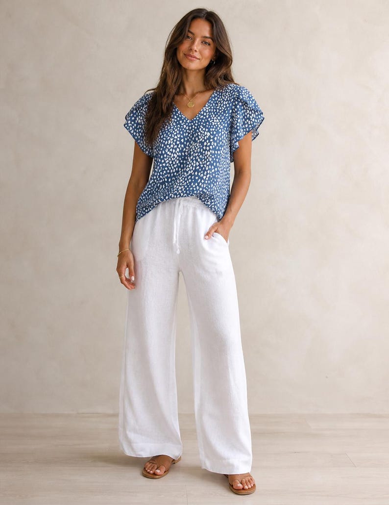 May include: A blue V-neck blouse with a white speckled pattern and flutter sleeves paired with white wide-leg trousers featuring a drawstring waist and side pockets. The outfit is completed with brown open-toe sandals.