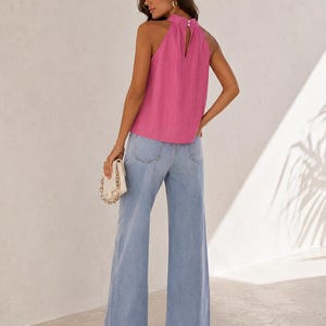 May include: A bright pink sleeveless halter top featuring a high neckline with two small white buttons at the back closure, paired with light blue wide-leg denim jeans. A small white handbag with a gold chain strap is held in one hand.
