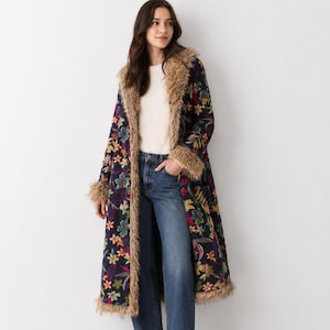 May include: A long, dark navy blue coat featuring a vibrant, multi-coloured floral pattern throughout. The coat is accented with thick, tan-coloured faux fur trim along the collar, front opening, and sleeve cuffs, paired with blue denim jeans.