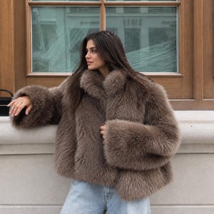 May include: A brown faux fur jacket with a high collar and long, wide sleeves. The jacket features a thick, textured pile and is worn over light blue denim jeans. The garment has a boxy, cropped silhouette that reaches the waistline.