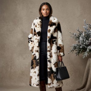 May include: A long faux fur coat with a cow print pattern in shades of brown, black, and white. The coat is worn over a black turtleneck and pants. A black handbag is held in the hand. The coat has a wide collar.