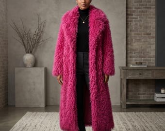 May include: A long, vibrant pink faux fur coat. The coat has a wide collar and a full-length design. The model is wearing a black turtleneck and black pants. The background features a neutral-toned brick wall and a rug.